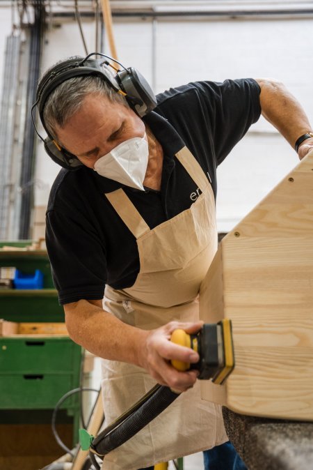 A man wearing ear defenders, a mask, and an apron uses a power sander on a wooden surface in the ercol factory