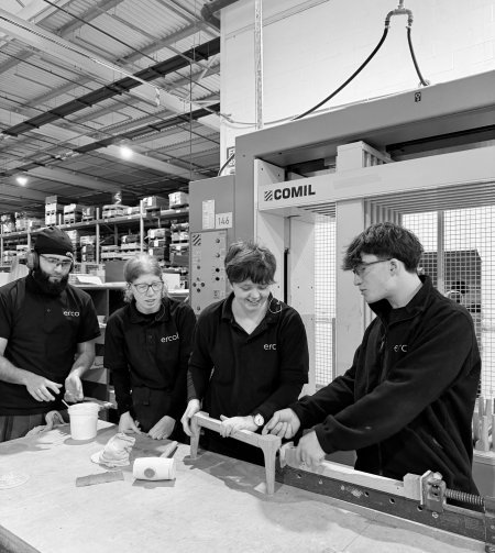 Four people in ercol uniforms assemble wooden parts at a table in a factory, with shelves and machinery visible in the background